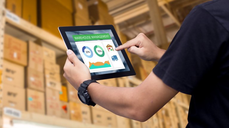 Close up of hands holding tablet in front of stacks of boxes in a warehouse.
