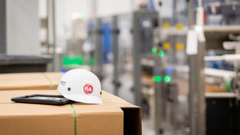 A white hard hat with red Rockwell Automation logo sits on a stack of corrugated flattened boxes with modern factory equipment in the background. Great Lakes Cheese factory on Thursday, May 27, 2021 in Hiram, OH