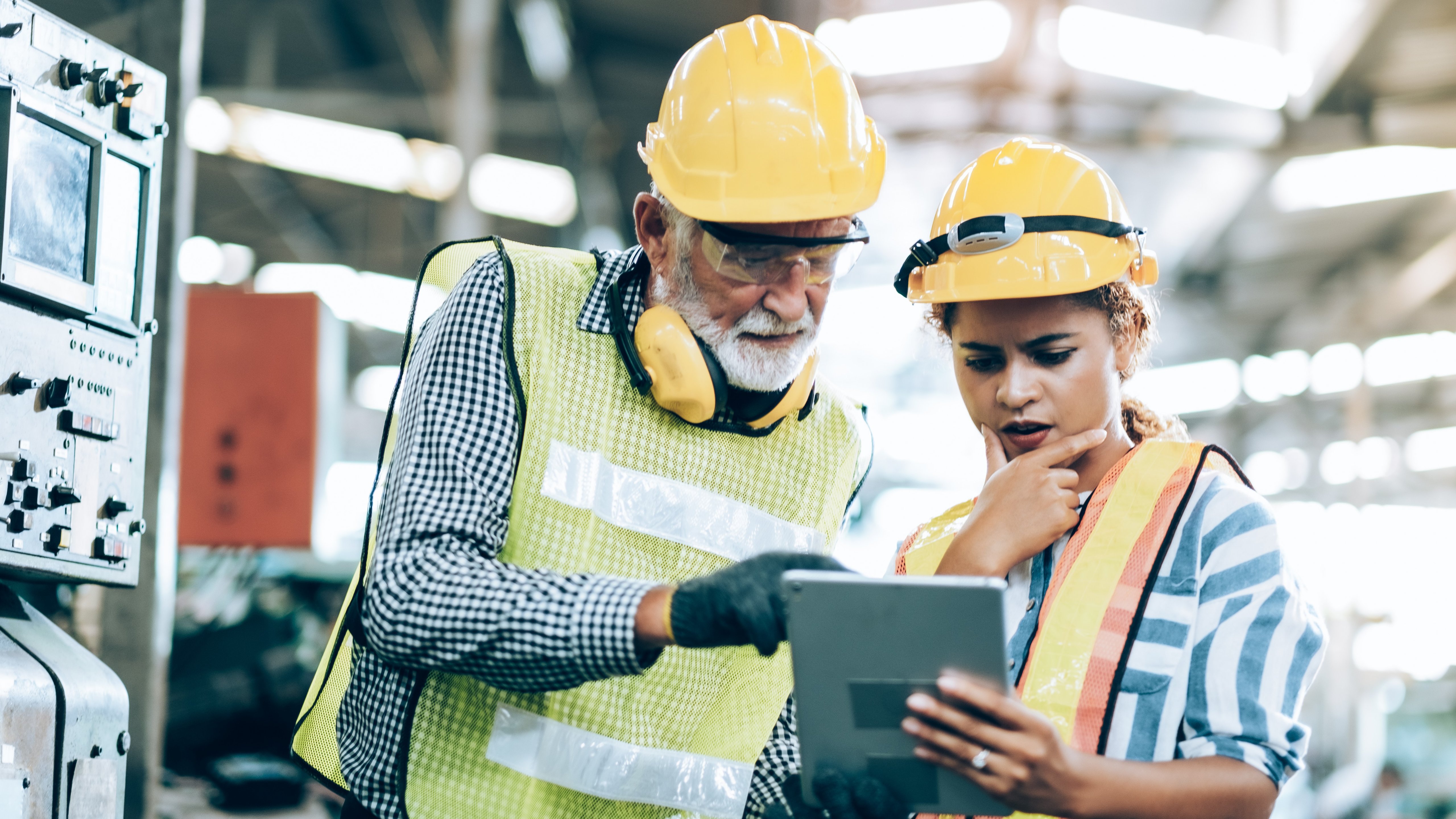 Portrait of Industrial Engineers in Hard Hats and Vests Working at a Heavy Industry Manufacturing Factory. Employees are Looking at a Tablet in an Indoor Factory.