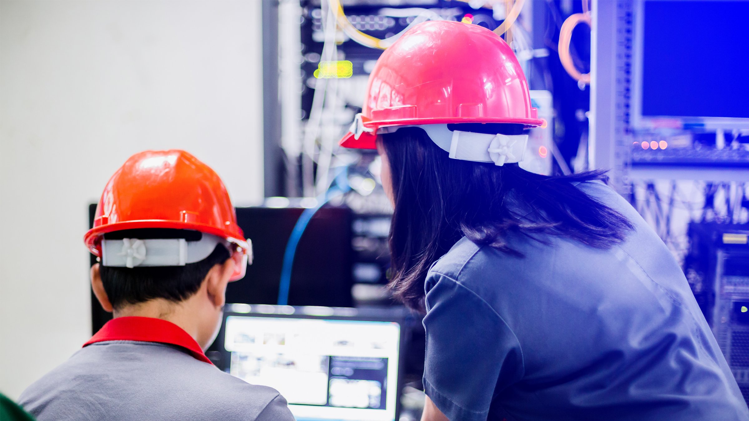 Back view of two people wearing red safety helmets leaning over a computer