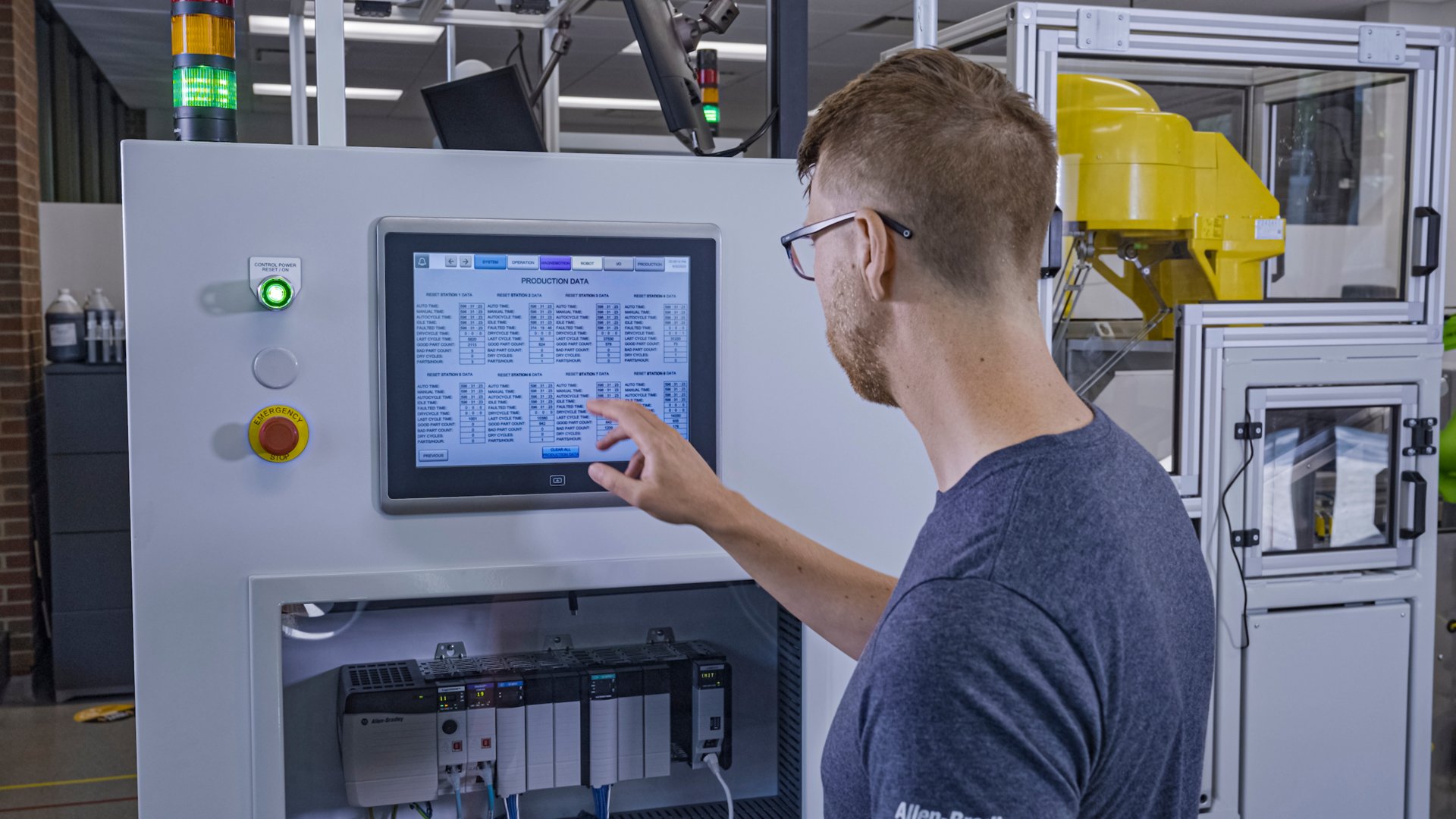 A male robot system operator reaches towards an HMI screen as a yellow robot runs in the background.