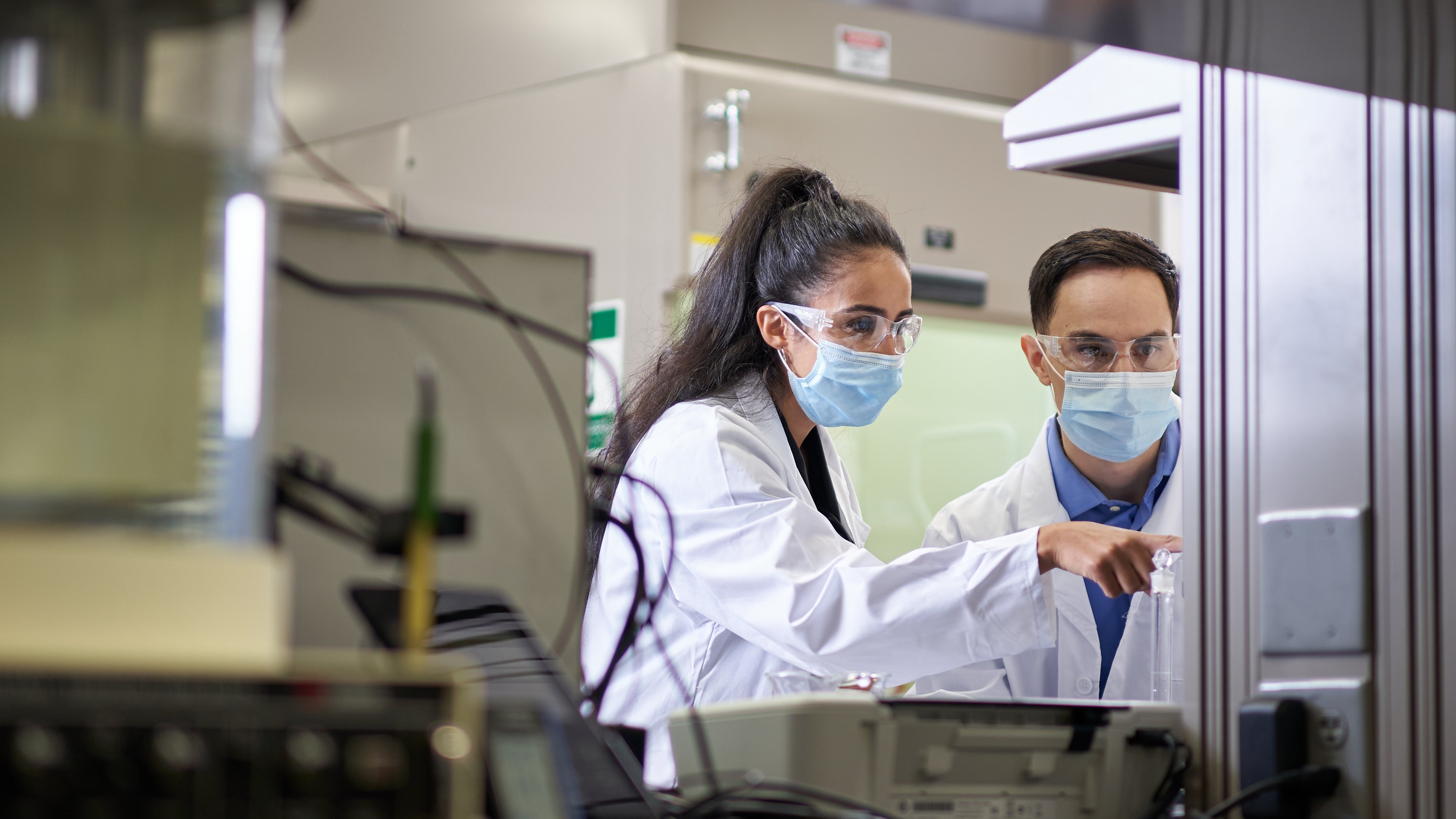 Life Sciences workers wearing lab coats andmasks and protective glasses while pointing at machine