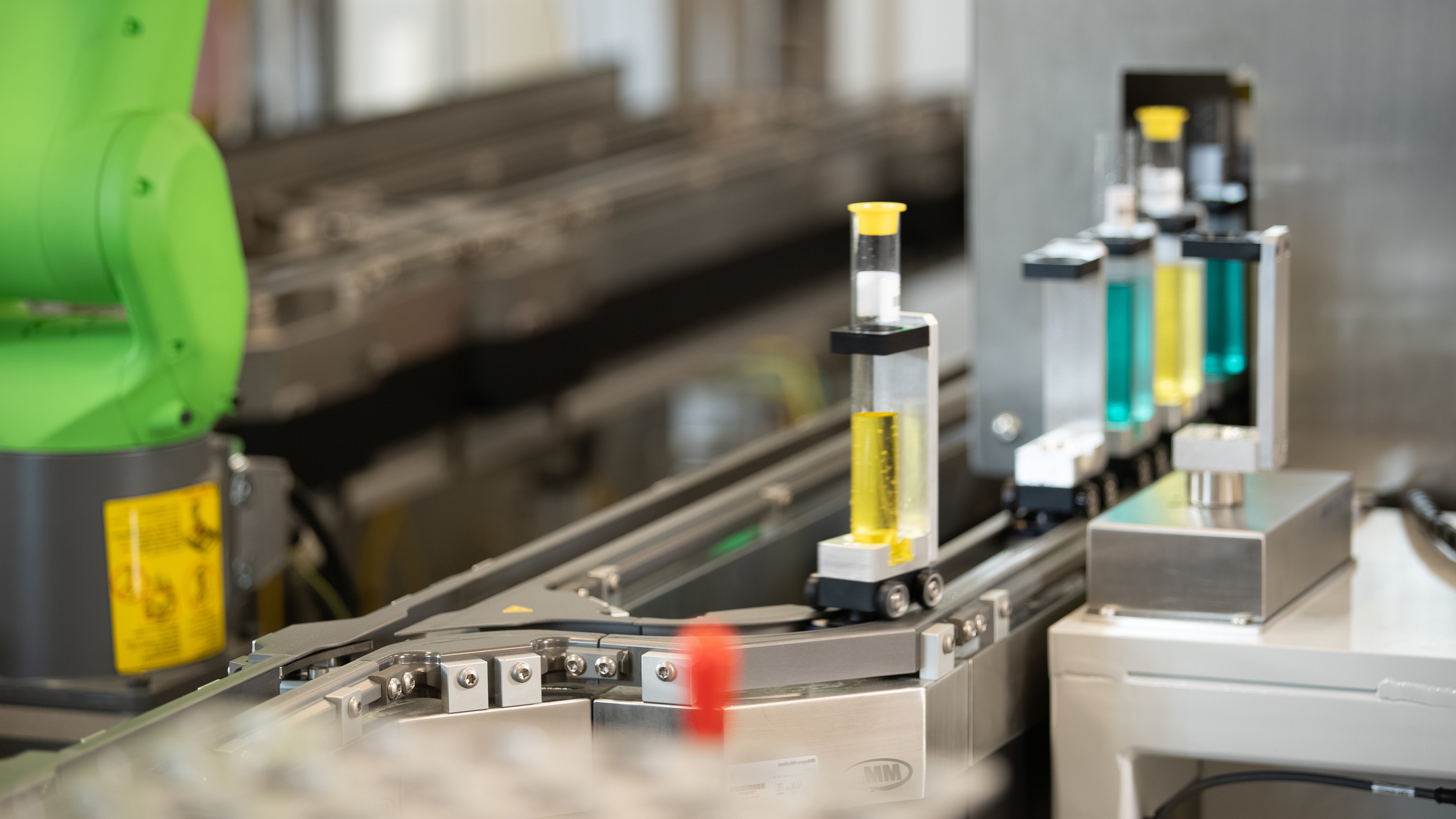 A MagneMover LITE Intelligent Conveyor System with a mover carrying a test tube filled with yellow liquid and a robot and other movers in the background.