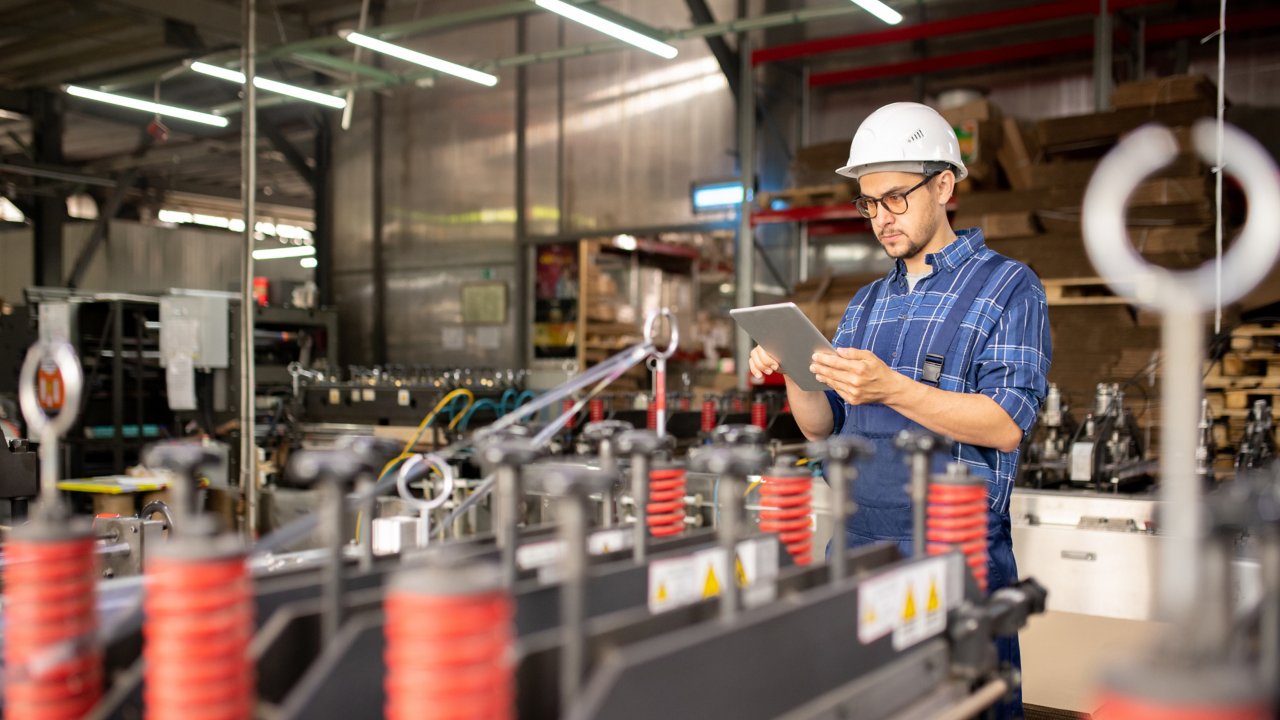 Young male manufacturing engineer on plant floor wearing hard hat and holding tablet. Manufacturing engineer holding tablet looking for information on industrial cybersecurity.