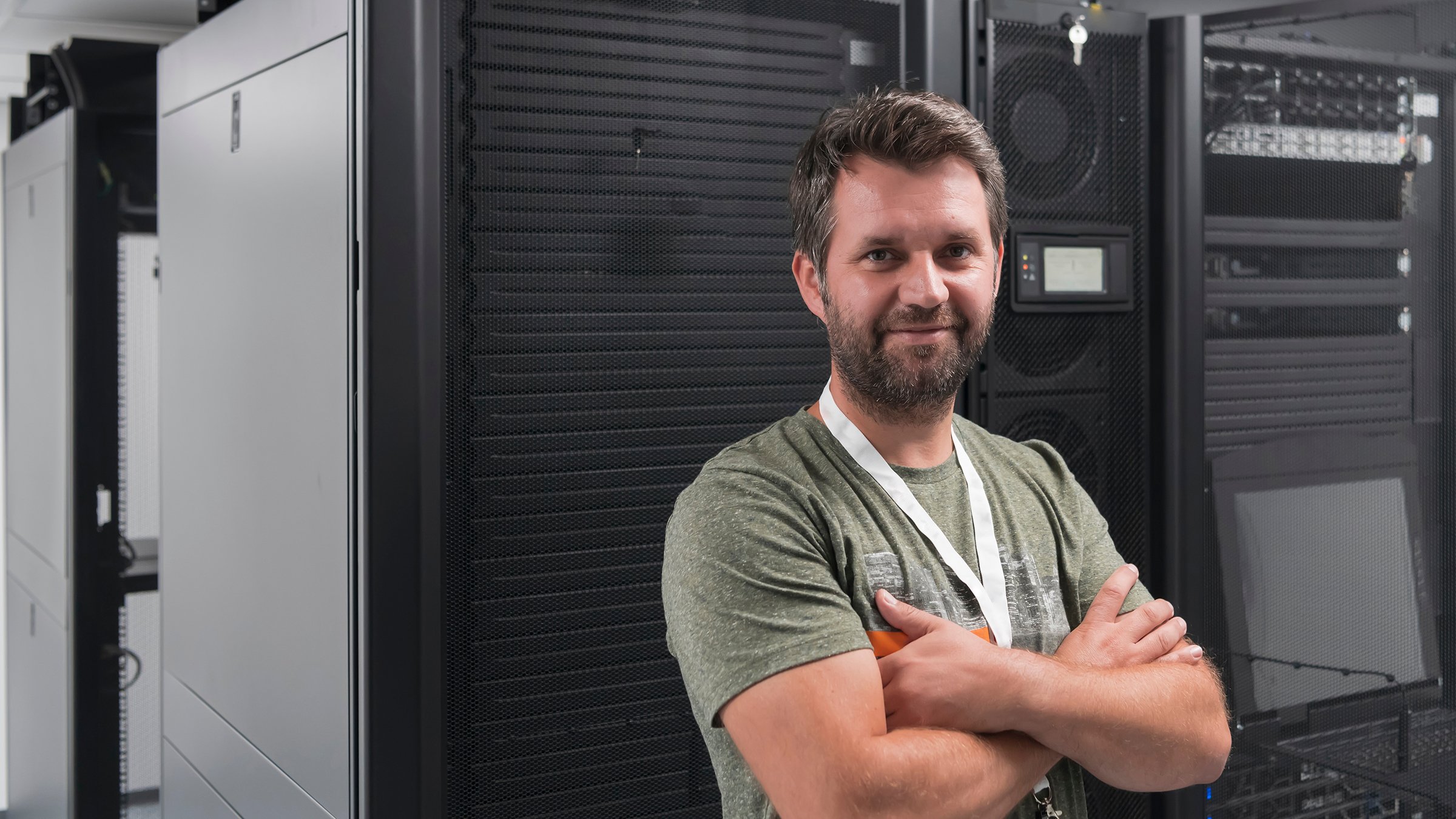 Male engineer wearing t-shirt and lanyard standing in server room in front of industrial data center with arms crossed.