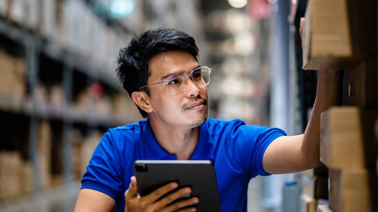 A male worker in blue shirt consults storeroom shelves for critical parts inventory at manufacturing facility while holding digital tablet.