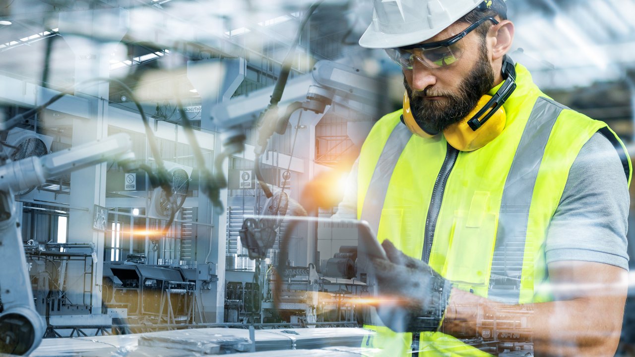 A bearded male wearing protective equipement looks at a tablet on the manufacturing floor in a factory.