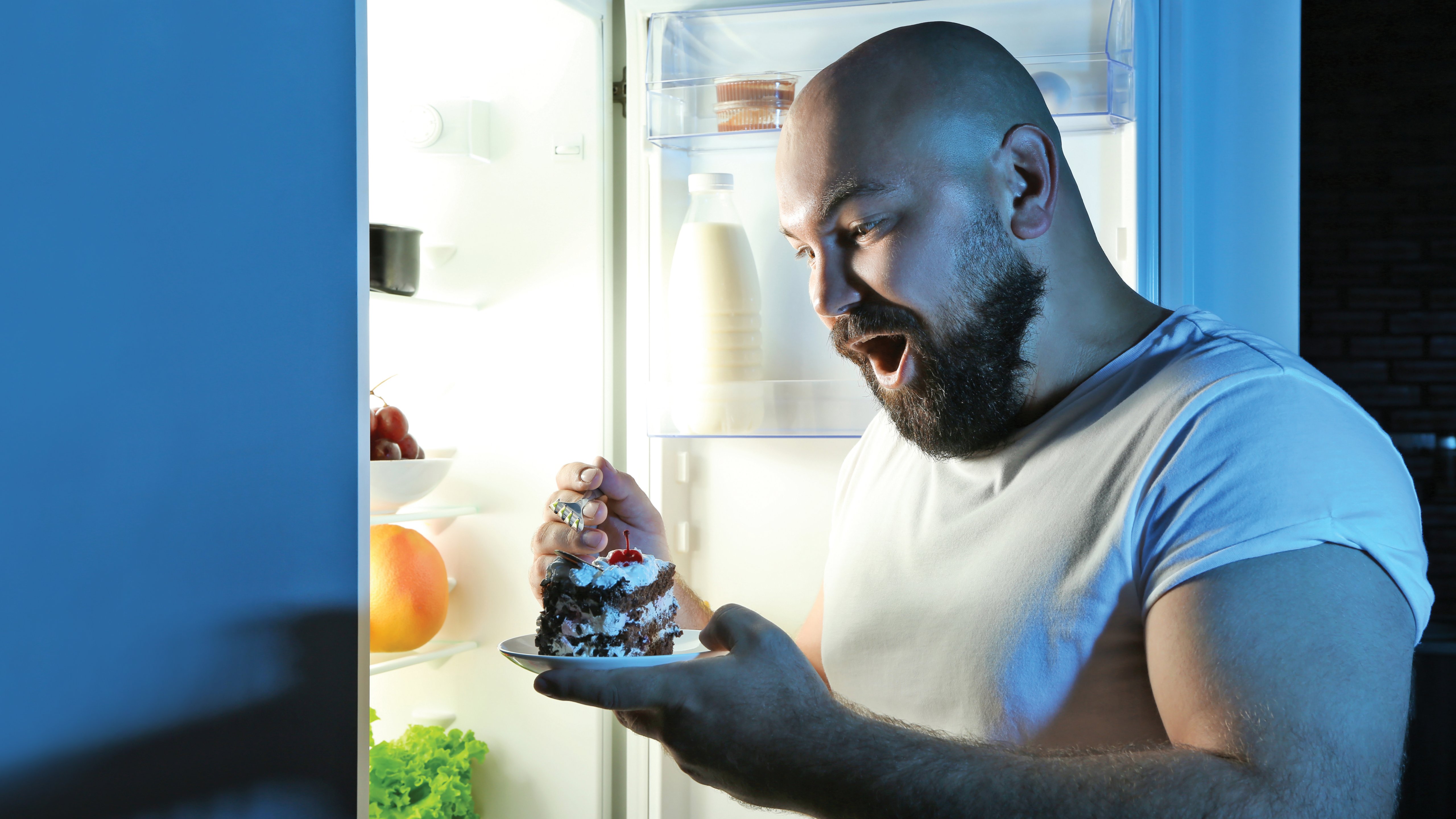 Man eating a cake at night with refrigerator door open