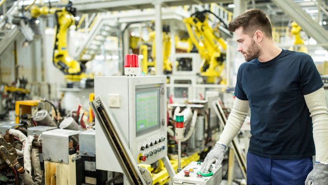 A male worker in blue shirt and pants uses an HMI to operate manufacturing equipment in a modern factory with yellow multi-axis robots in the background. Safety components like the light curtain, Emergency Stops, and indicator lights are recommended in risk assessment to safeguard personnel from harm and protect machinery and facilities.