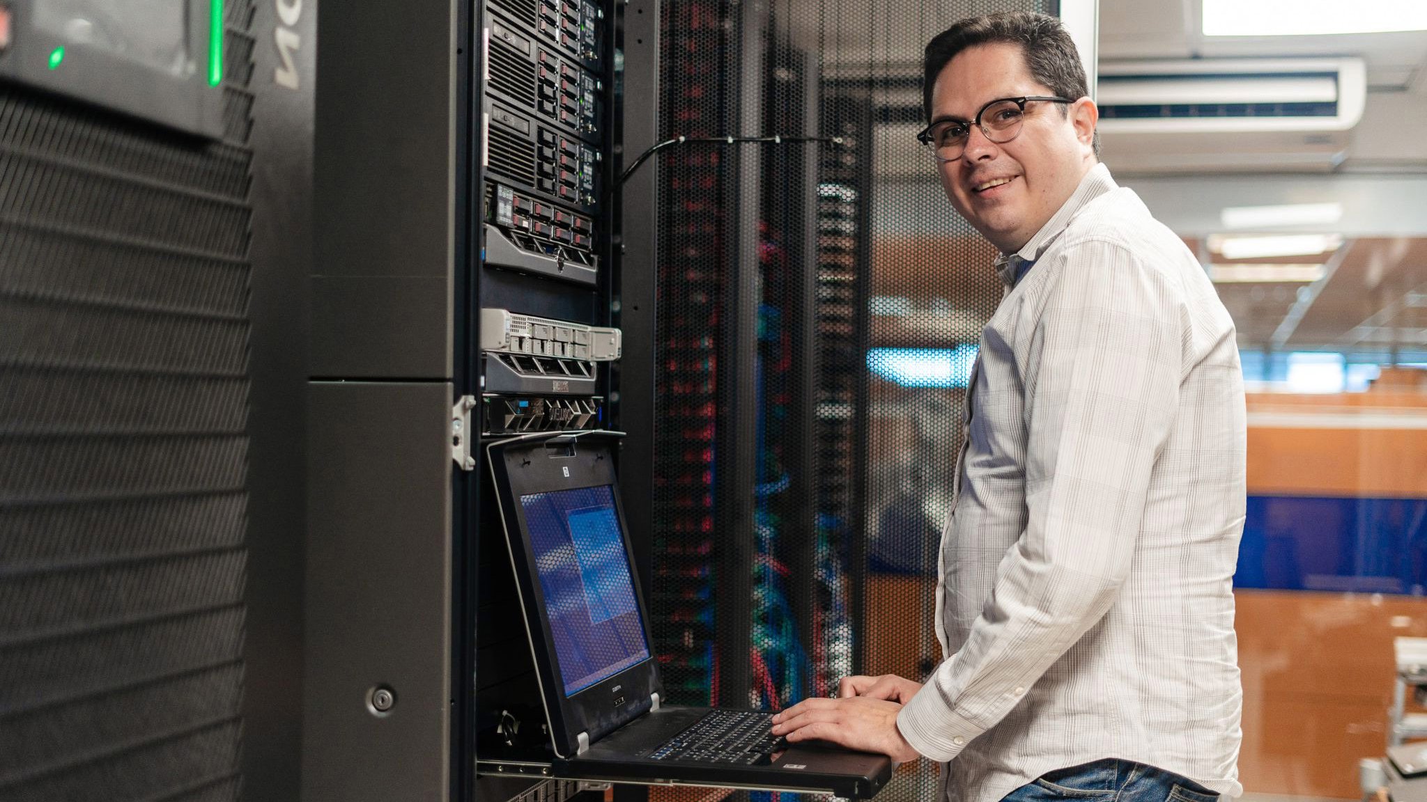 Man with brown hear wearing business casual clothing and glasses stands in front computerin Church & Dwight manufacturing industrial data center.