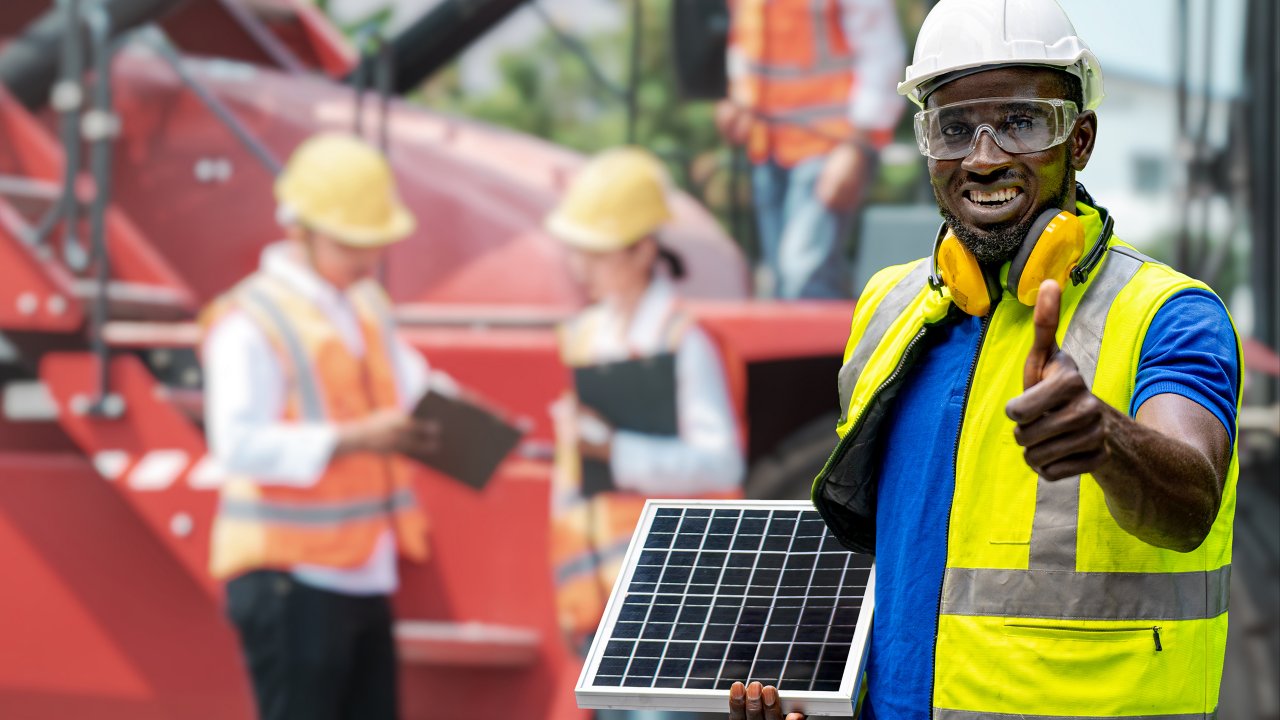 Man in PPE standing and giving thumbs up and holding solar cell panel. Workers meeting in front of heavy machine in background.