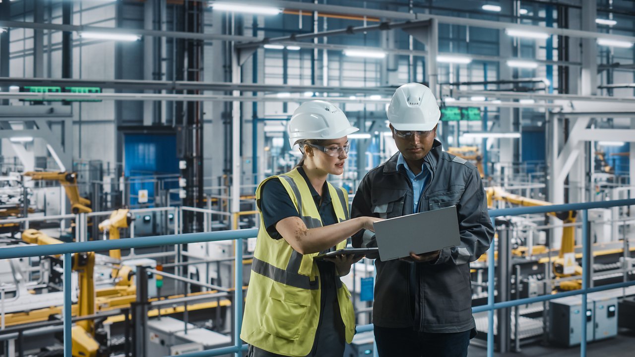 Una trabajadora y un trabajador revisan algo en un computador portátil en medio de una planta industrial