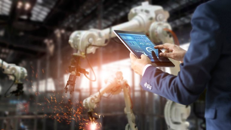 Un homme utilisant une tablette contrôle une machine automatisée munie de bras robotisés dans une usine industrielle intelligente sur un logiciel de système de surveillance en temps réel.