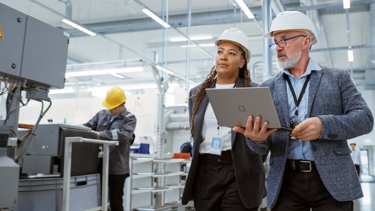 Two Professional Heavy Industry Engineers Wearing Hard Hats at Factory. Walking and Discussing Industrial Machine Facility, Working on Laptop. African American Manager and Technician at Work.