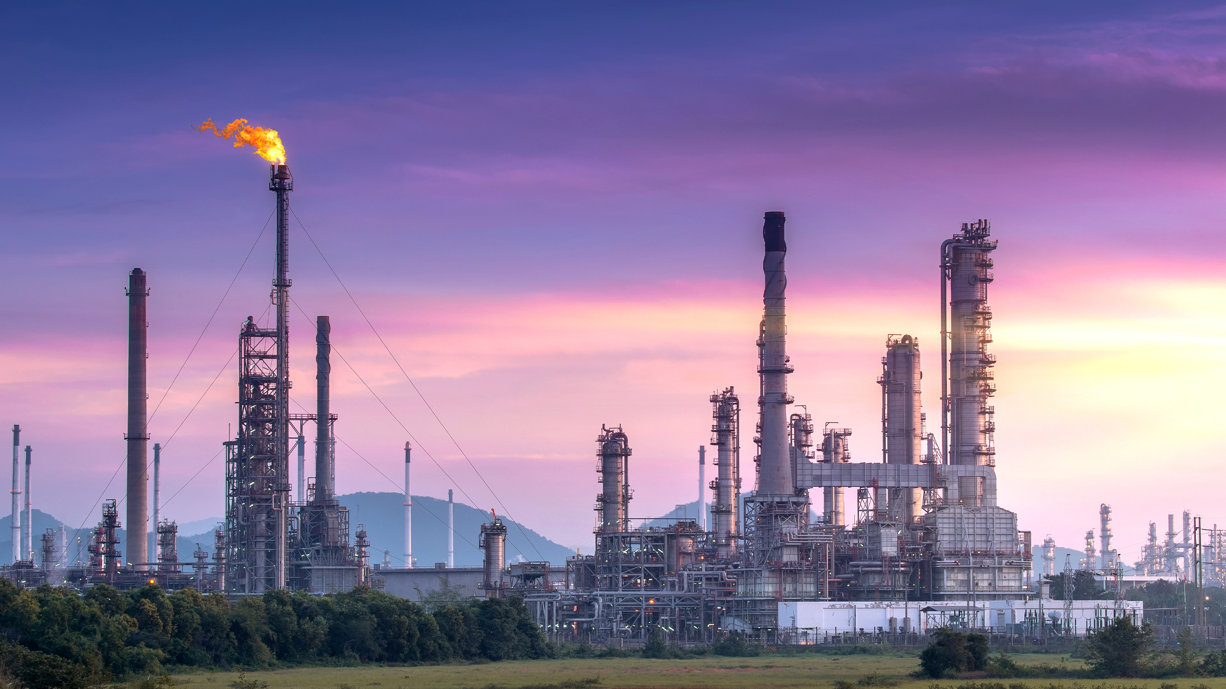 Refinería de petróleo recortada contra una puesta de sol, con humo saliendo de sus chimeneas hacia el cielo colorido