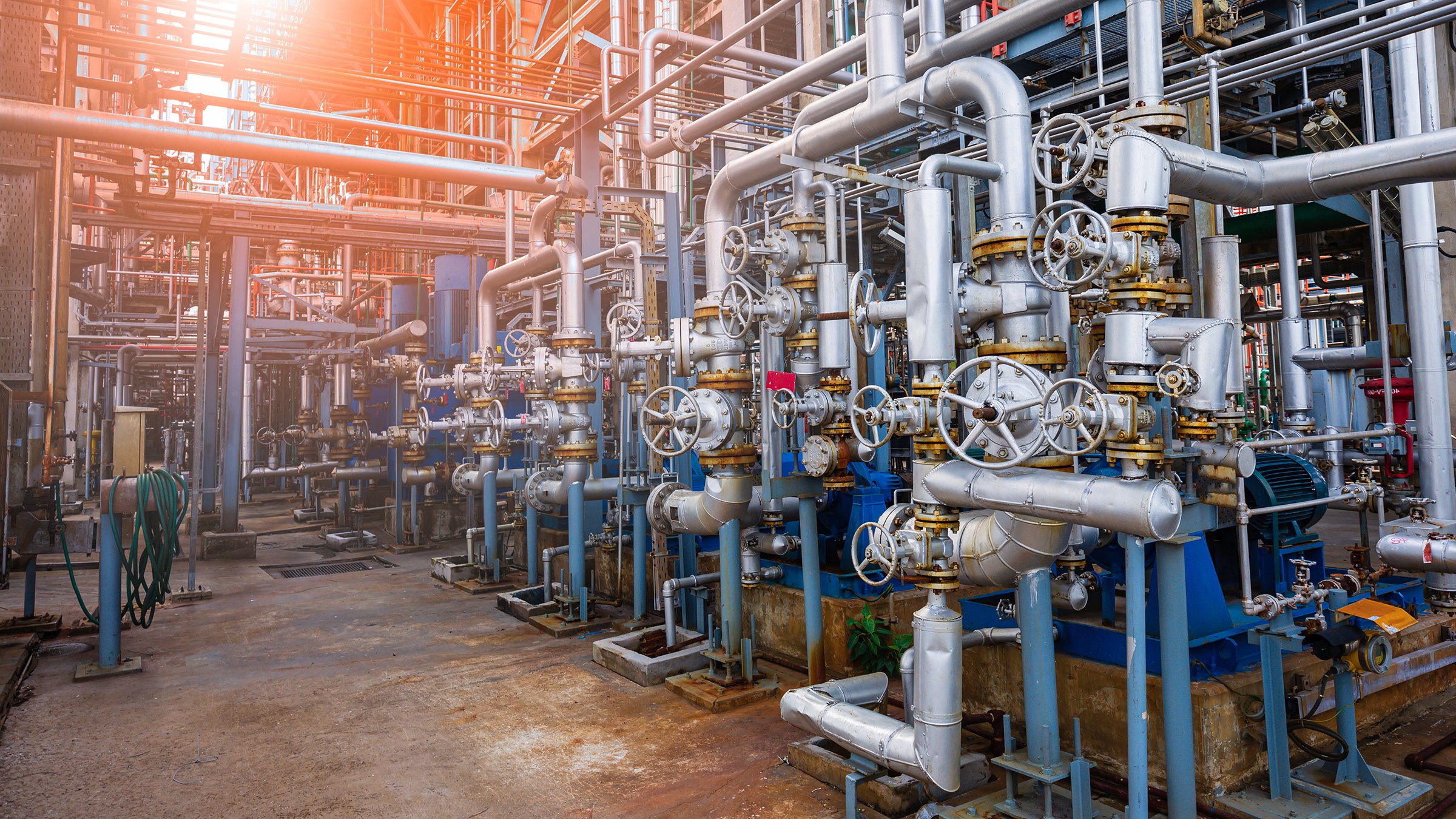 Uma diversidade de grandes tubos, conectores, motores e rodas industriais na cor prata, entrecruzados em vários níveis de uma refinaria de petróleo e gás ao ar livre.