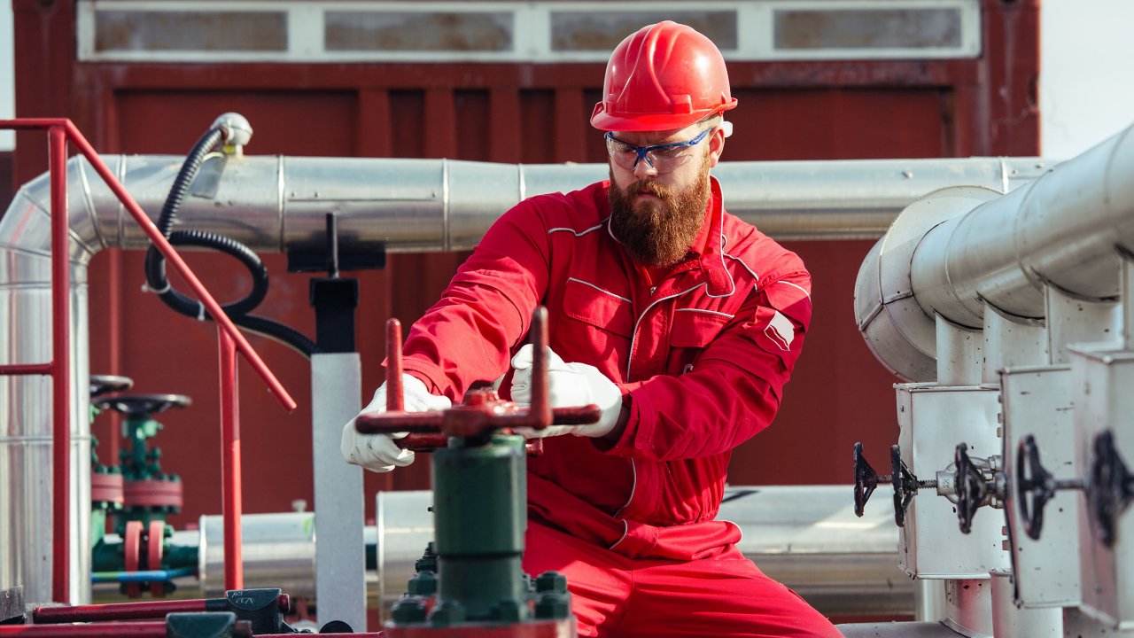 Un lavoratore della stazione di pompaggio di una raffineria di petrolio con un caschetto rosso e un’uniforme apre le valvole delle condutture. 