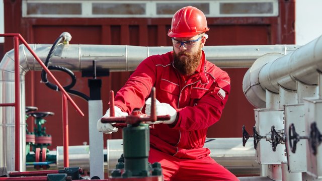 Un trabajador de una estación de bombeo de una refinería petrolera con casco rojo y uniforme abre válvulas de tuberías. 