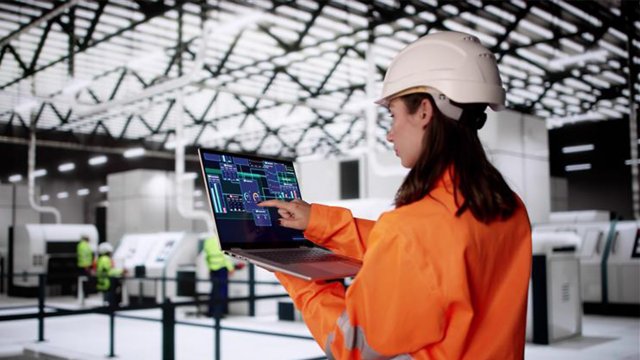 Female engineer in orange hazard suit wearing white hard hat holding a laptop computer on the plant floor analyzing manufacturing equipment on the plant floor for NIS2 OT security compliance