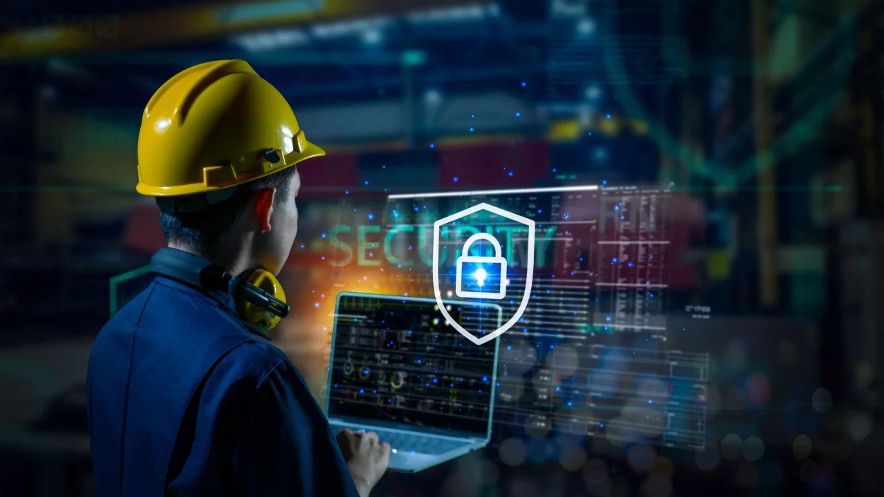 Engineer wearing yellow hard hat and noise cancelling headphones holds a laptop on the manufacturing plant floor with OT cybersecurity imagery concept covering background.