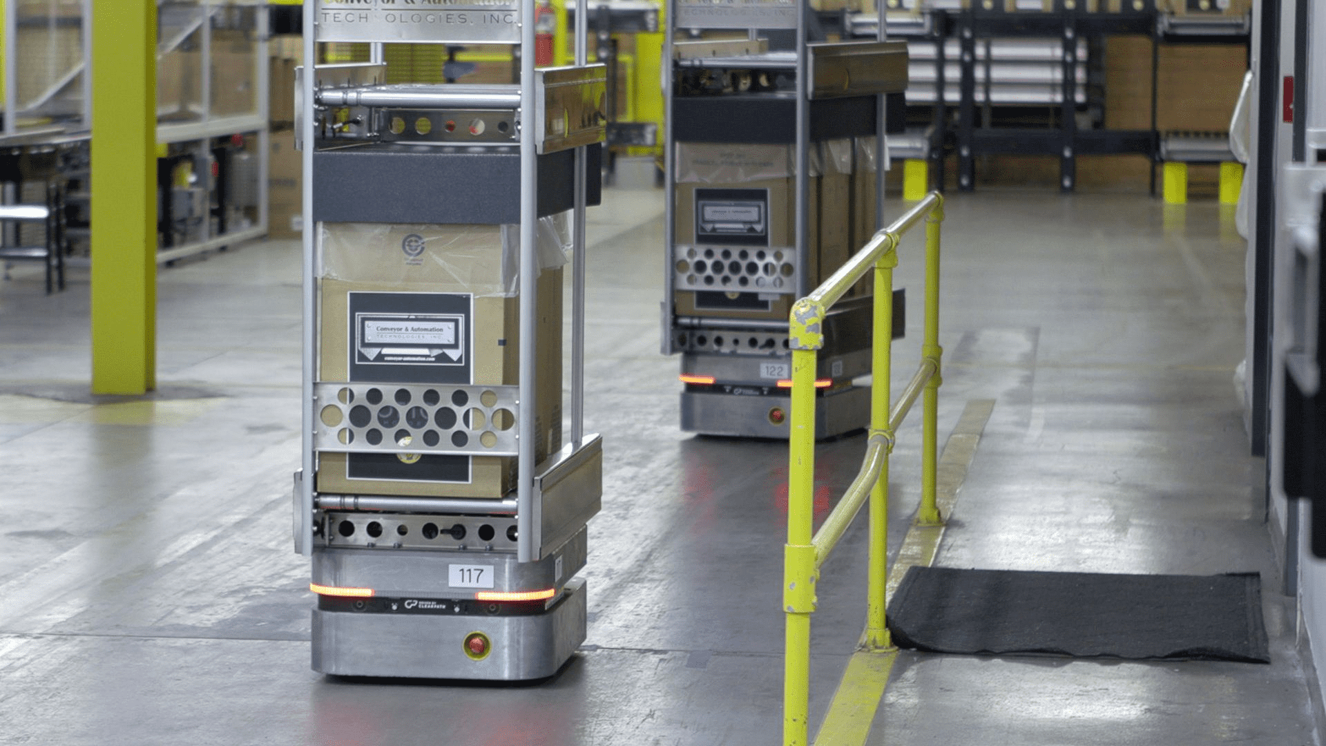 Two silver autonomous mobile robots move carts filled with products on a factory floor