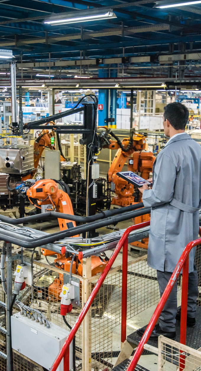 manufacturing facility interior with robotic arms and a man in a suit holding a tablet