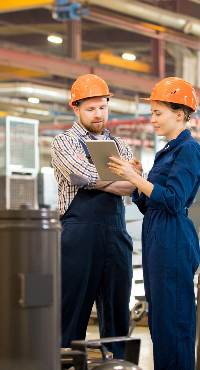 Two manufacturing workers in orange hard hats discussing work in a factory setting