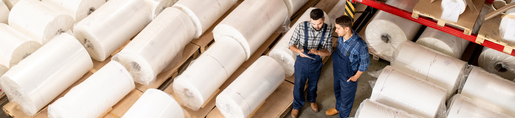 Workers discussing in a warehouse with large rolls of foam stacked on shelves