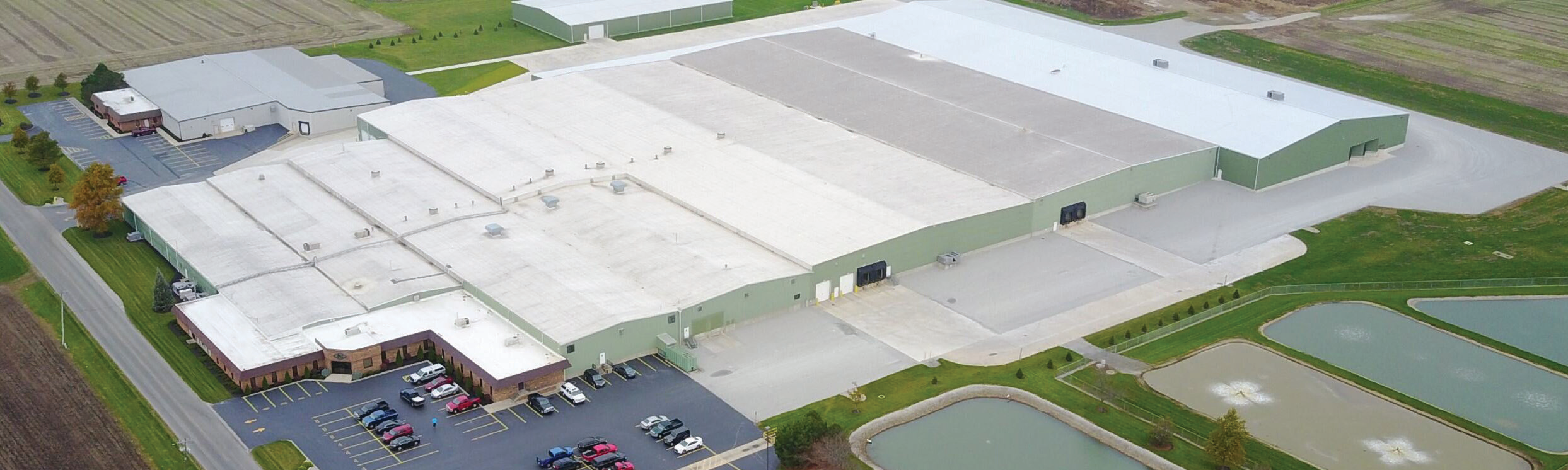 aerial view of a manufacturing facility exterior with parking lot and surrounding green fields