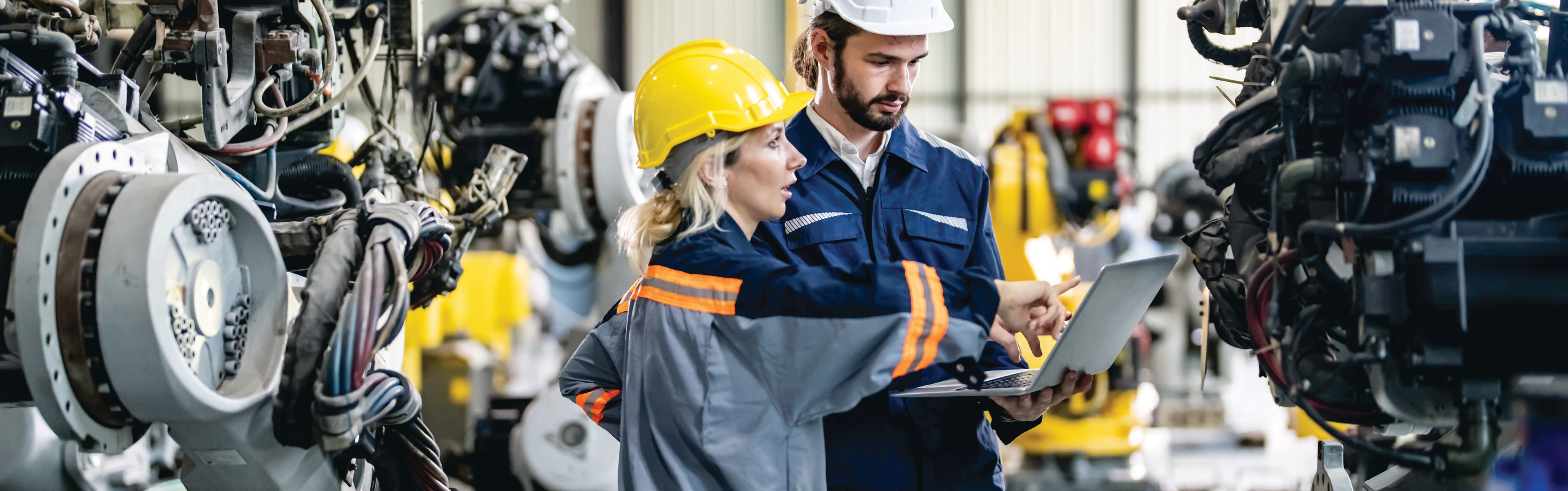 manufacturing facility interior with two workers in safety gear using a laptop among machinery