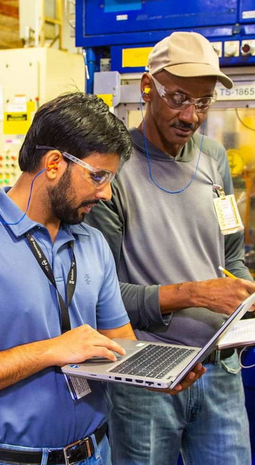 Two workers in a manufacturing facility, one using a laptop and the other holding a clipboard