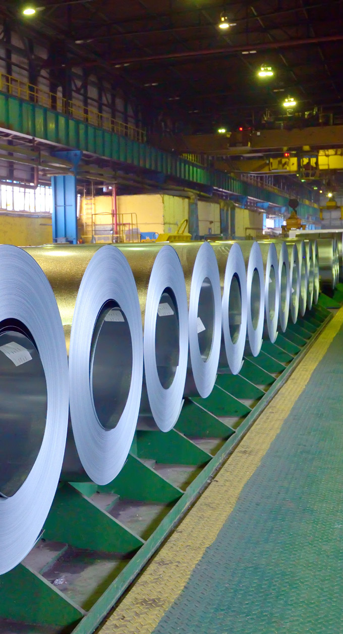 manufacturing facility interior with large rolls of metal sheet lined up on green stands under bright overhead lights