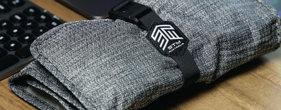 Folded gray fabric with STM Australia logo patch on black strap, placed on wooden surface with keyboard partially visible