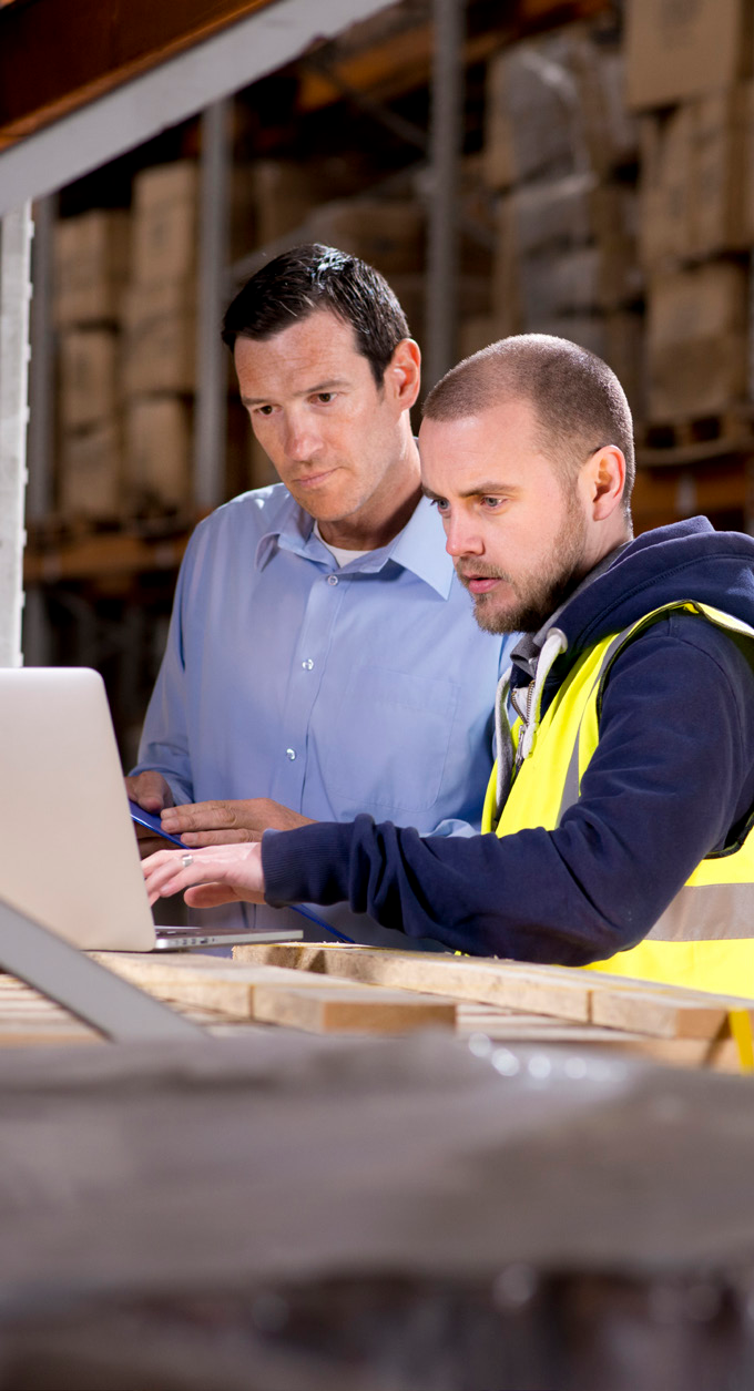 Two workers in a manufacturing facility using a laptop, wearing safety vests
