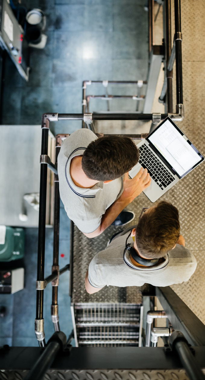 overhead photo of two people working on laptops in a manufacturing facility with visible pipes and equipment
