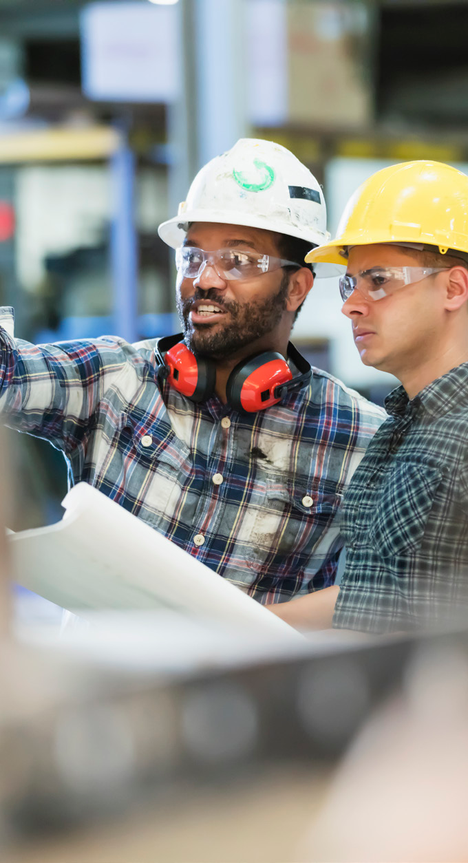 Two workers in hard hats and safety glasses reviewing a document in a manufacturing facility