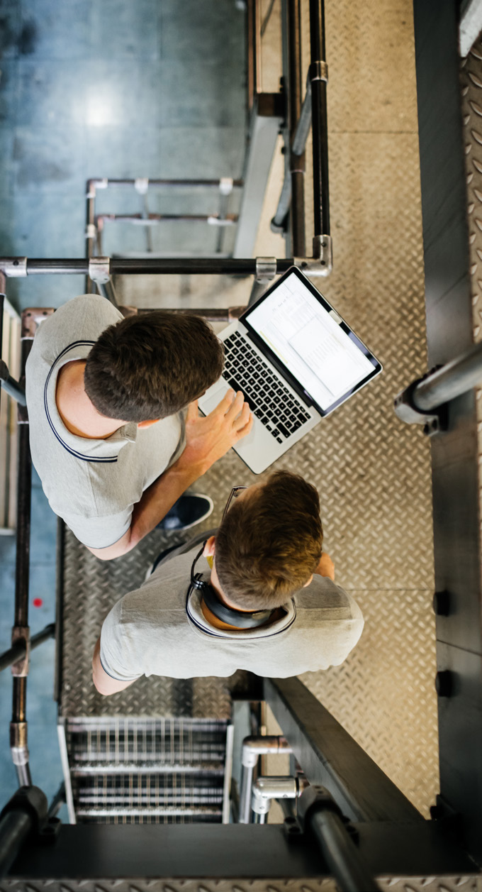 Overhead view of two workers standing near a laptop on a metal staircase in a manufacturing facility