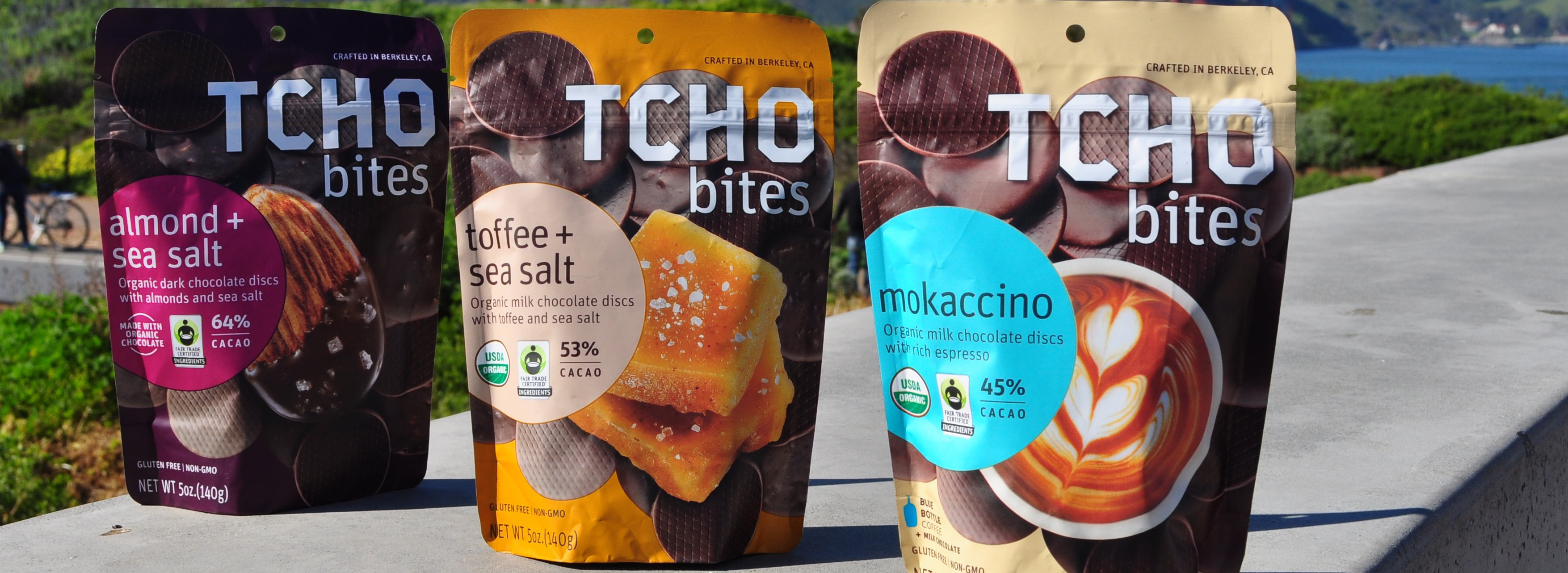 Three TCHO Bites chocolate product packages on a concrete surface outdoors