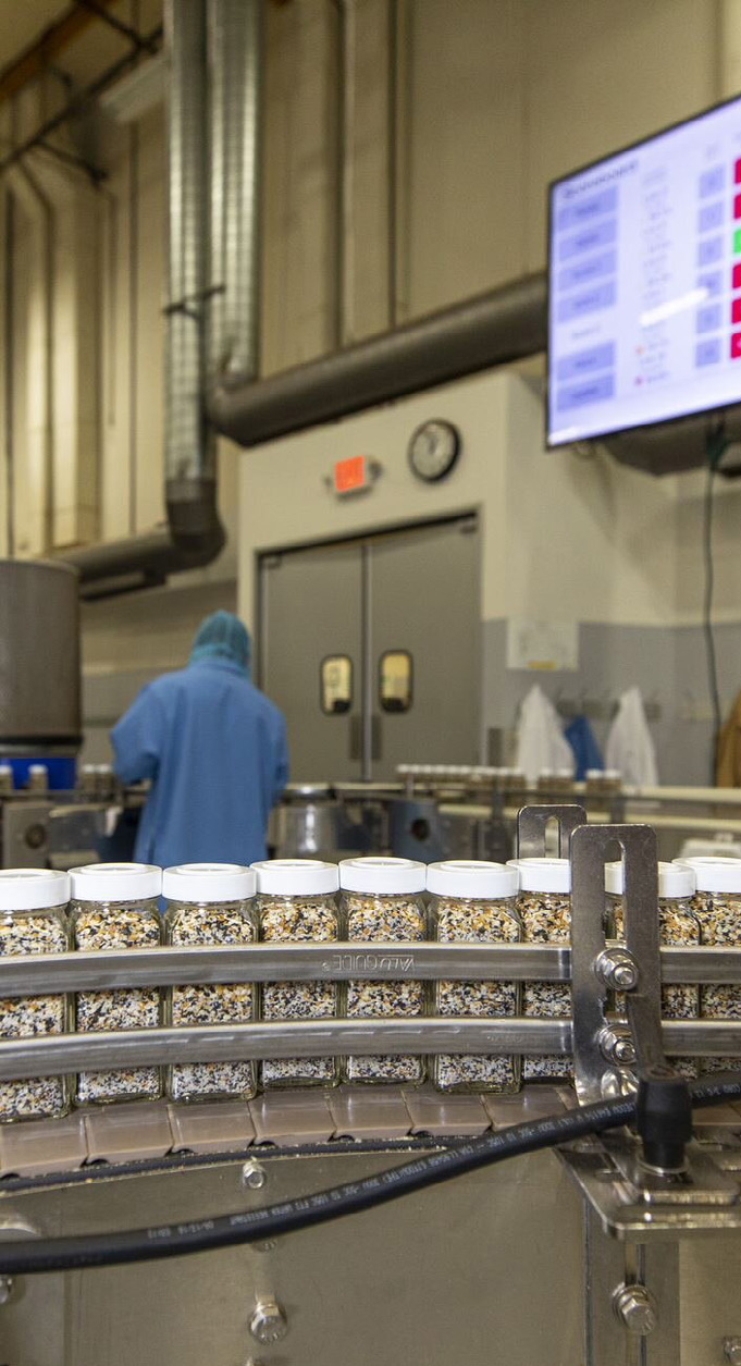 manufacturing facility interior photo with worker in blue coat and conveyor belt with spice containers