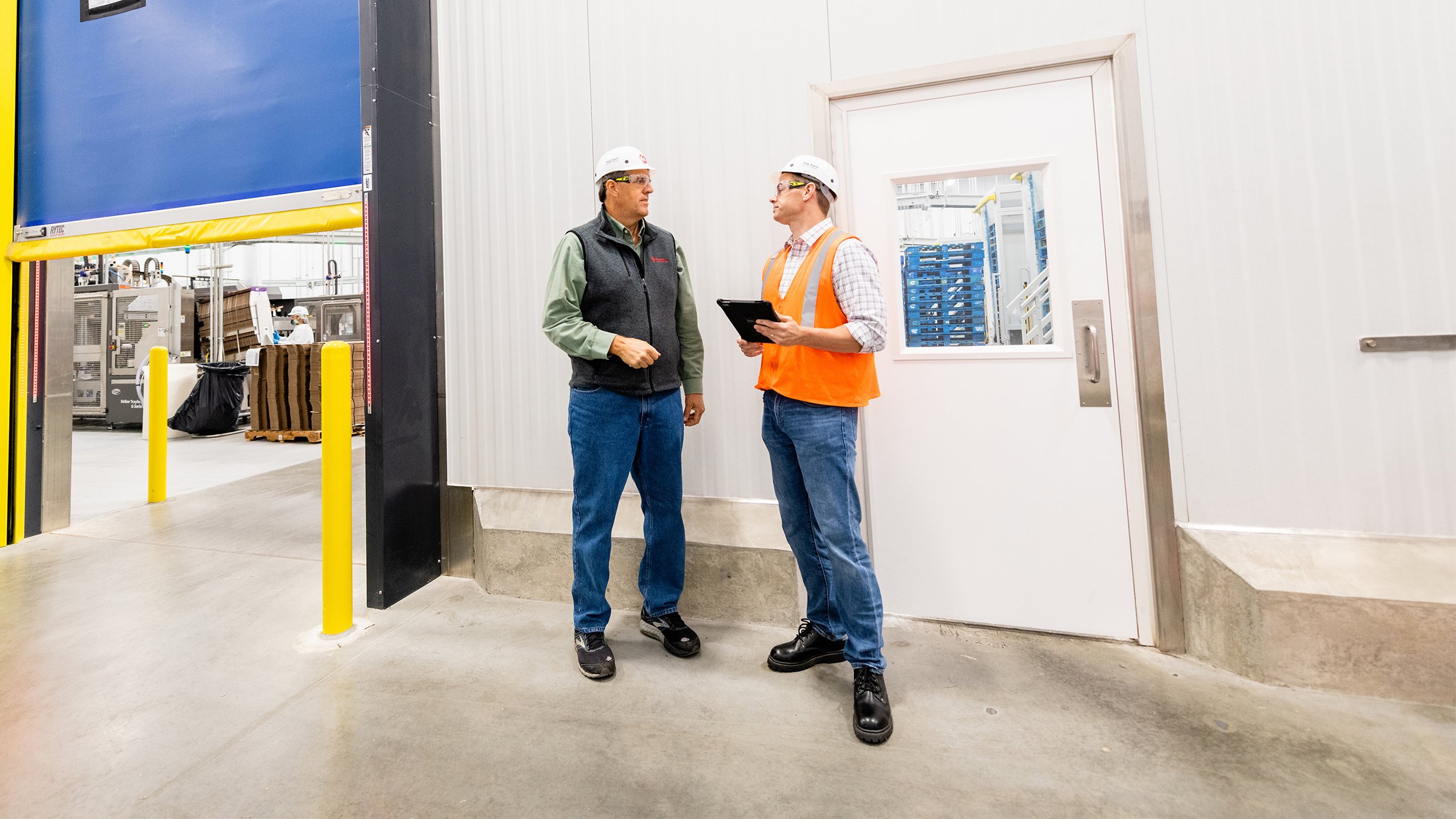 A male plant manager in high-visibility orange vest and white hard hat holds an electronic tablet while talking to a dedicated Rockwell Automation asset management professional about their repair and inventory reports in their Asset+ Managed service contract.