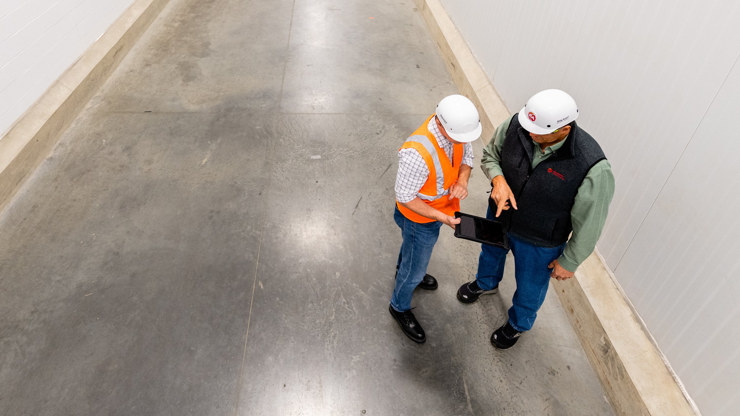 A male plant manager in high-visibility orange vest and white hard hat holds an electronic tablet while discussing their Asset+ Managed service contract with their dedicated asset management professional, also wearing a white hard hat and gray Rockwell Automation vest.