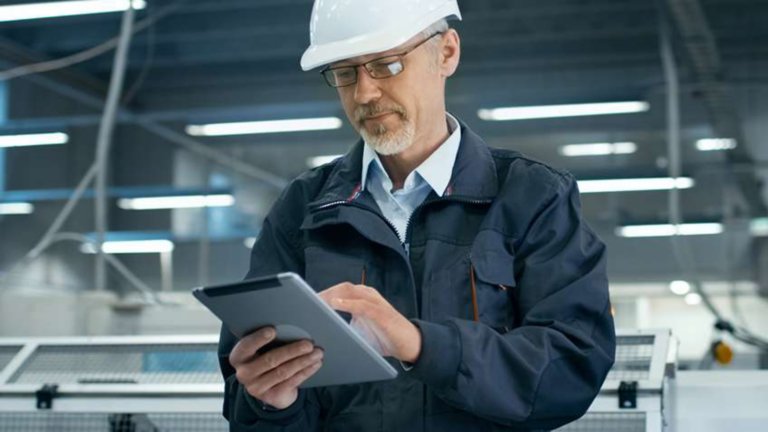 Male employee wearing a white hard hat in the factory typing data into a tablet