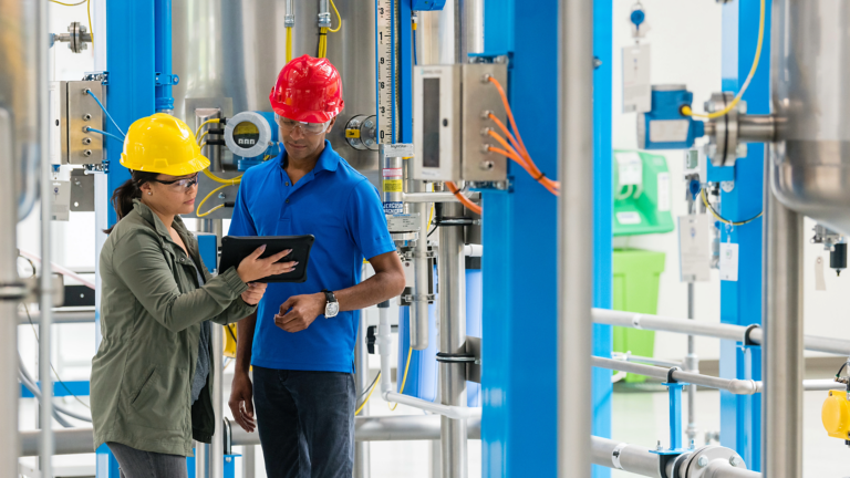 A female worker in yellow hard hat shows an ipad to a male safety manager in red hard hat as they discuss maintenance of their Rockwell Automation Plant PAx Distributed Control System in a factory environment