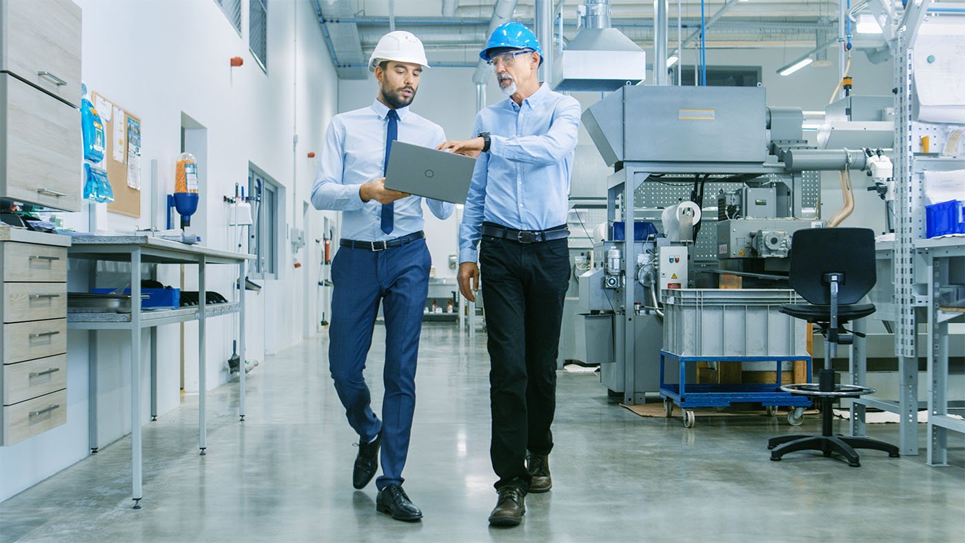 Male head of manufacturing holds laptop discussing product detains with chief engineer walking through modern factory.