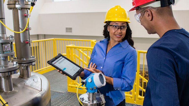 A female employee in yellow hard hat consults with a male application engineer to check that a production batch is functioning safely.