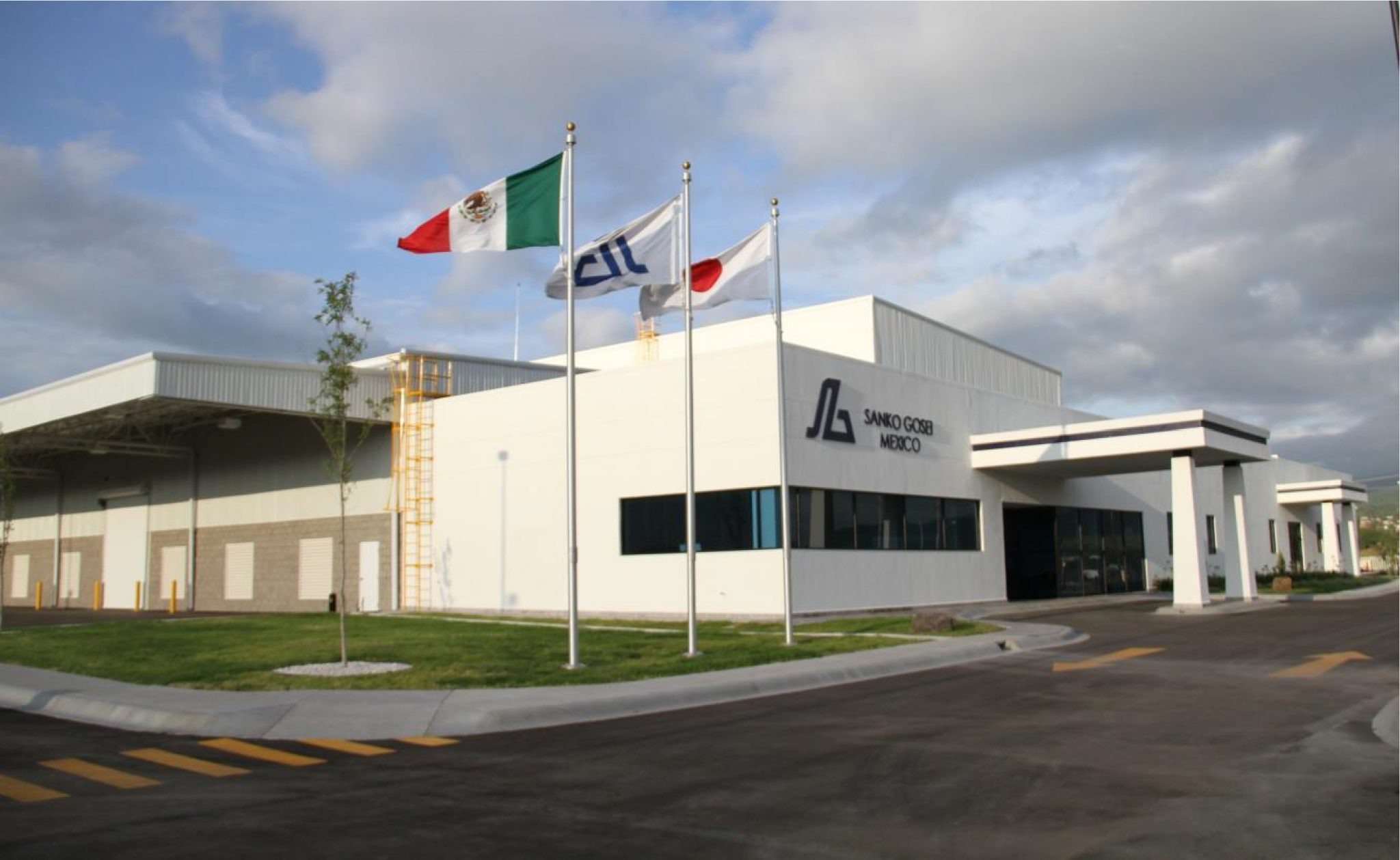 Exterior de un moderno edificio blanco de las instalaciones de Sanko Gosei México, destacando tres astas con las banderas de México, la empresa y Japón bajo un cielo parcialmente nublado