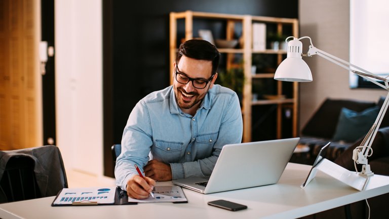 戴著眼鏡微笑的男子坐在辦公桌前使用筆記型電腦及書寫資料。