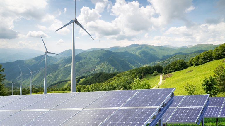 Solar panels with wind turbines against a mountain landscape against blue sky with clouds.