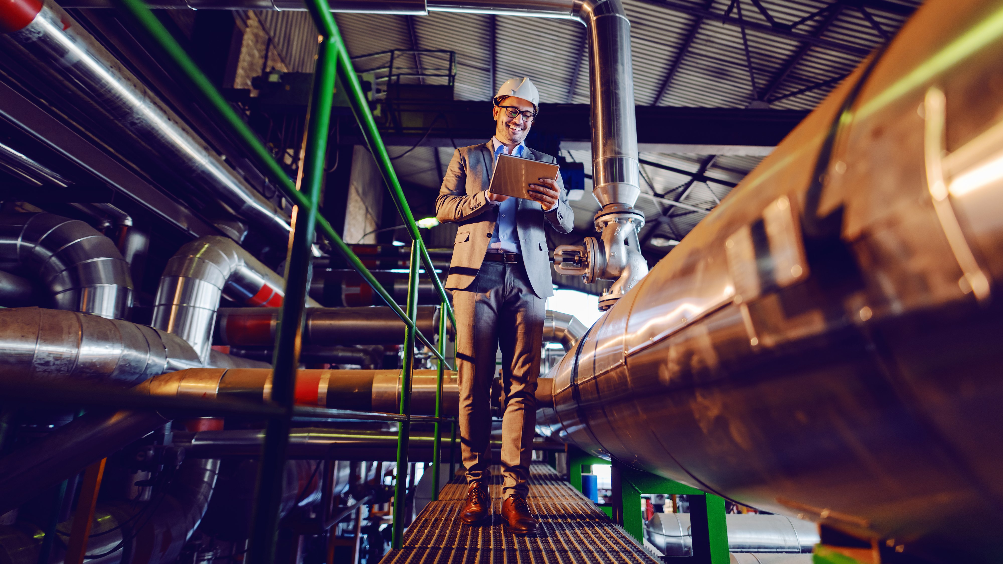 Handsome smiling caucasian supervisor in suit and with helmet on head using tablet while walking in power plant.