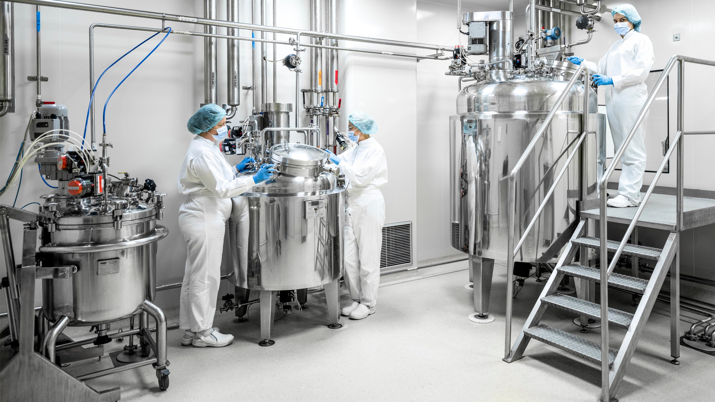 Three female workers in pharmaceutical industry wearing protective blue gloves, mask and cap and white suit seen standing by the machine that is the part of the medicaments production during the working hours in a pharmaceutical manufacturing.